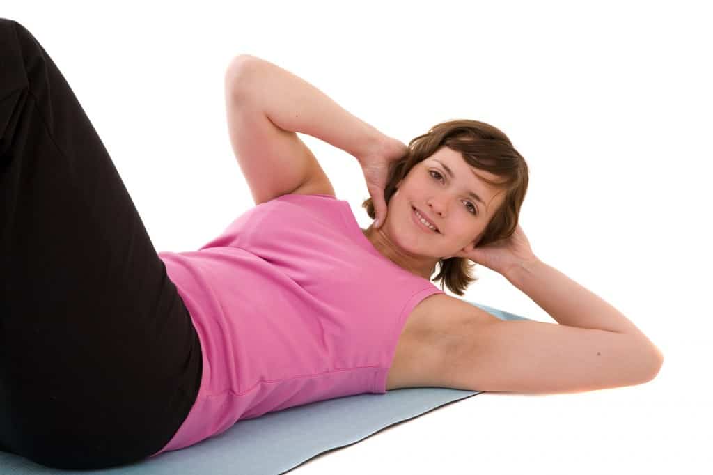 Young woman in a pink tank top and black pants lying on her back in sit-up position with her hands behind her head