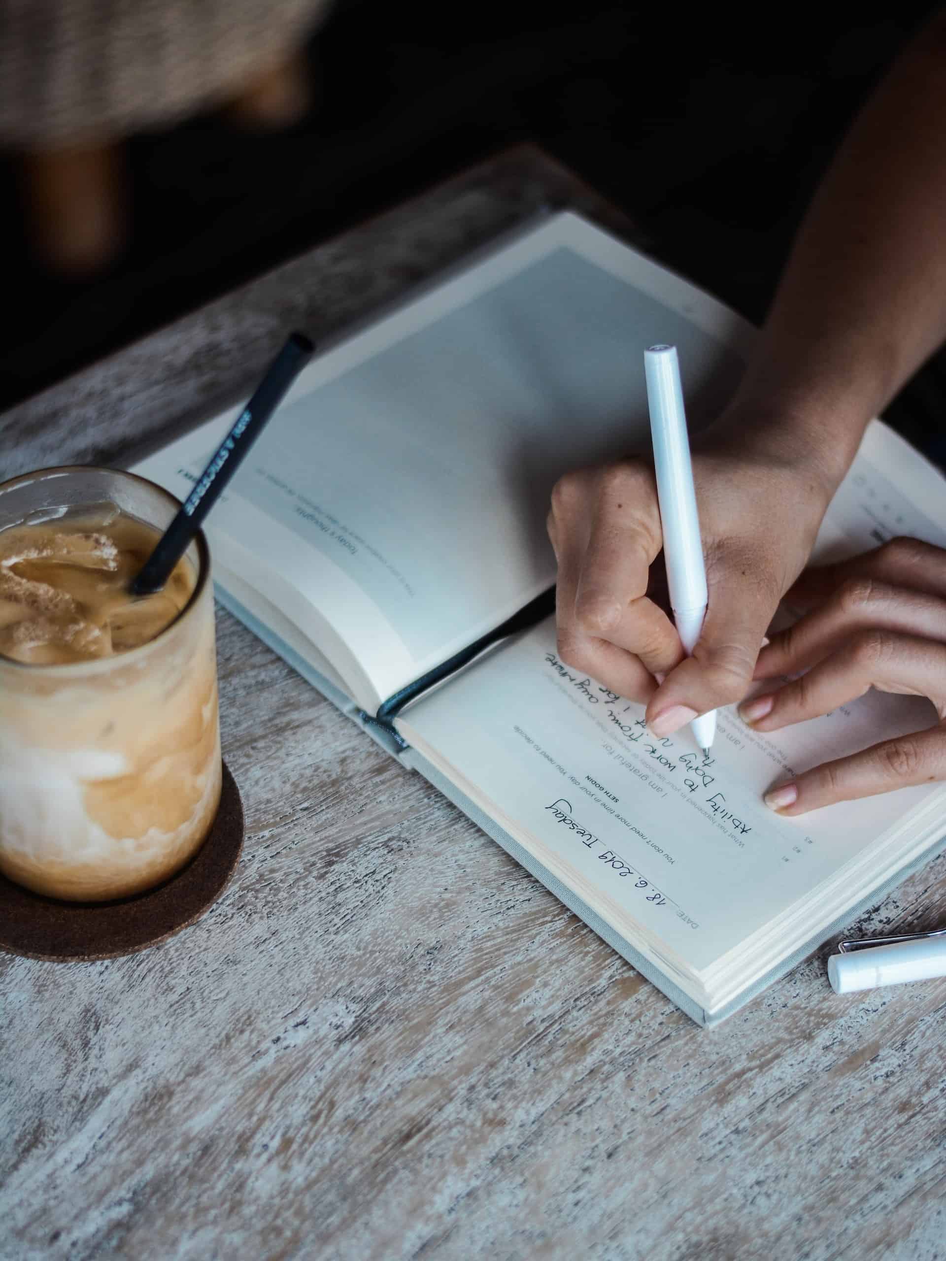 Photo of arms writing in a journal with iced latte on the desk in front