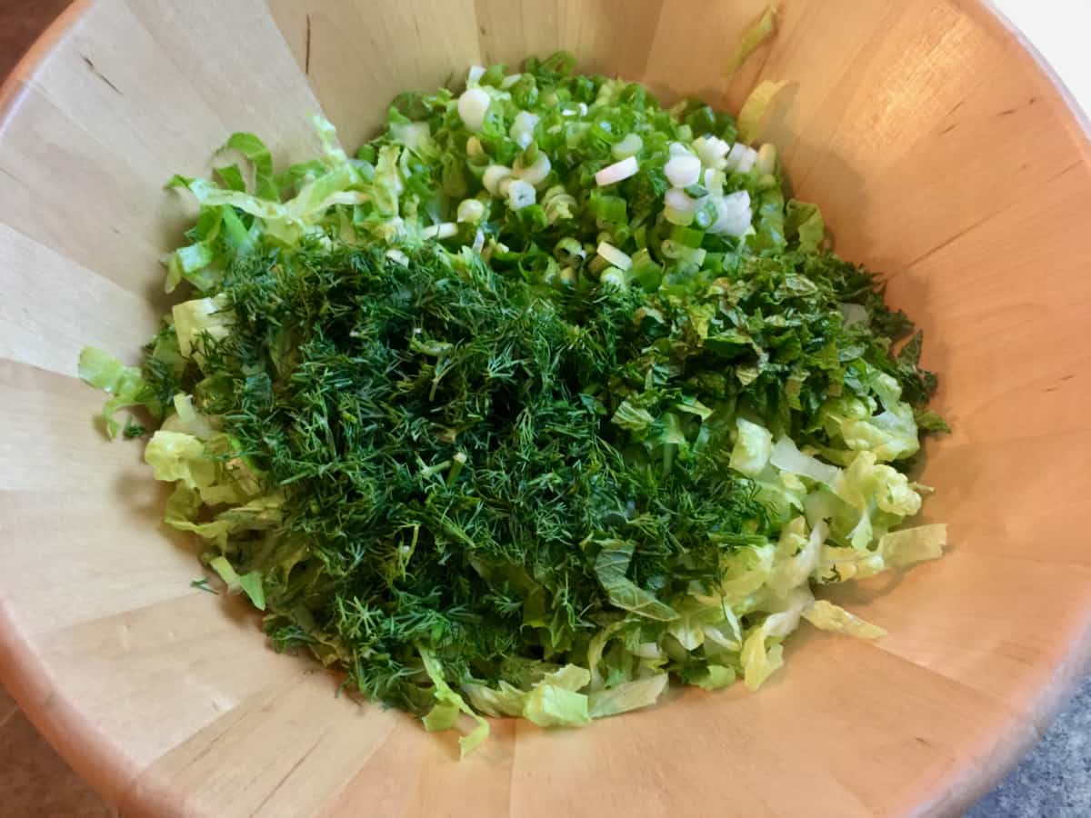 Combining shredded romaine lettuce, chopped green onion and dill in wooden salad bowl.