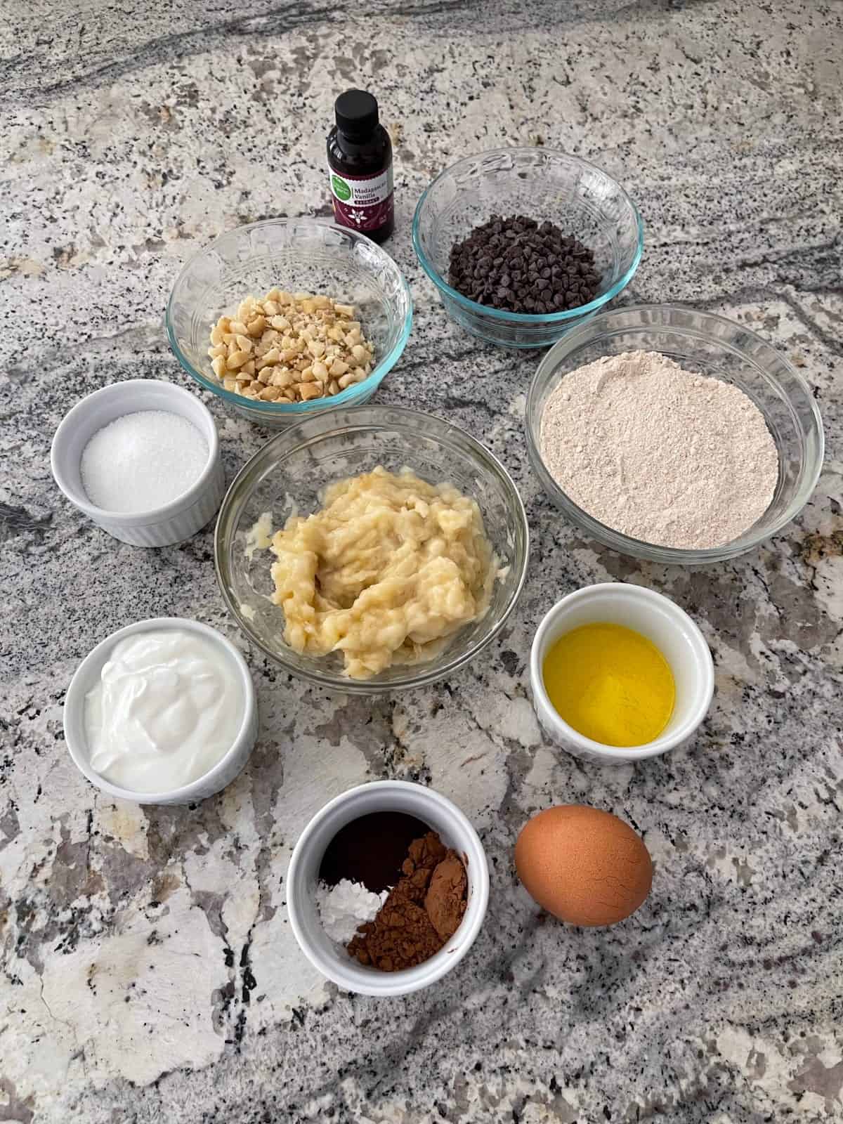 Ingredients including vanilla, macadamia nuts, mini chocolate chips, flour, mashed banana, Greek yogurt, sweetener, butter, egg and spices.
