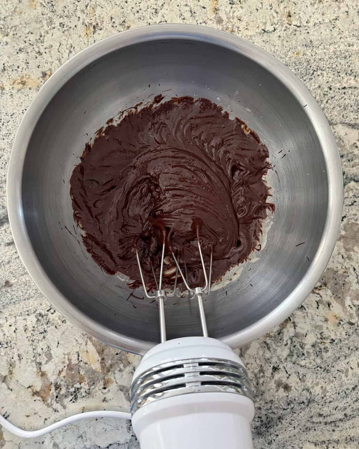 Blending eggs, sugar, vanilla extract and melted chocolate with handheld electric mixer in stainless mixing bowl.