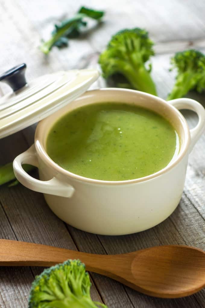 Small crock with creamy broccoli soup, a wooden spoon and broccoli florets scatter about