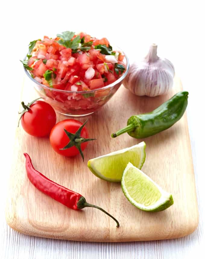 Pico de gallo salsa in glass bowl on wood cutting board with tomatoes, chili peppers, lime wedges and garlic bulb.