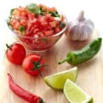 Fresh pico de gallo salsa in glass bowl with tomatoes, lime wedges, chili peppers and garlic on wooden cutting board.
