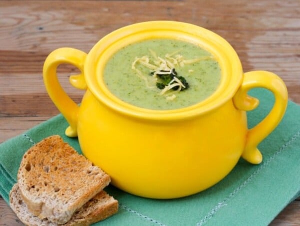 Cream of Broccoli Cheese Soup in a bright yellow crock sitting on a green placemat with toast alongside