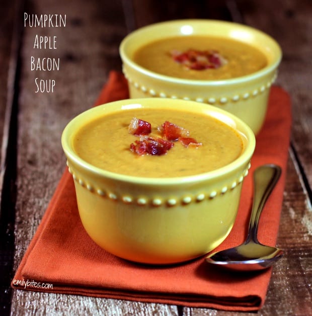 Two bowls of pumpkin soup topped with crumbled bacon on a wooden table with a spoon and orange napkin