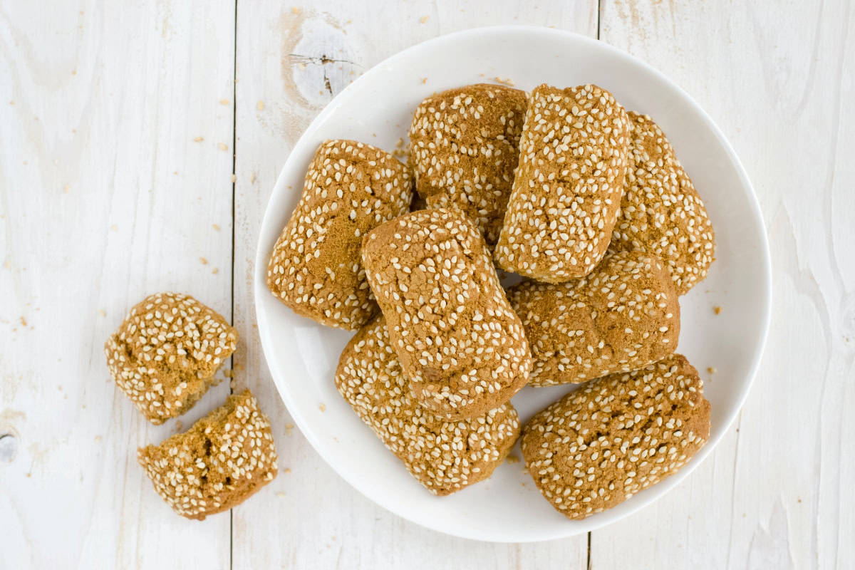 Italian sesame seed cookies stacked on a white plate on a white table with one cookie broken in half sitting next to the plate.