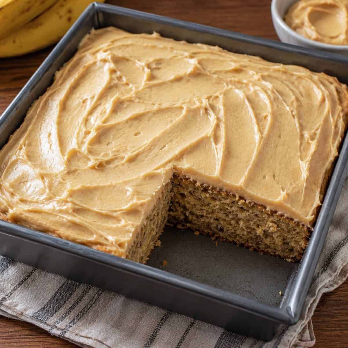 Banana cake with peanut butter frosting in a square baking pan with a piece cut out.