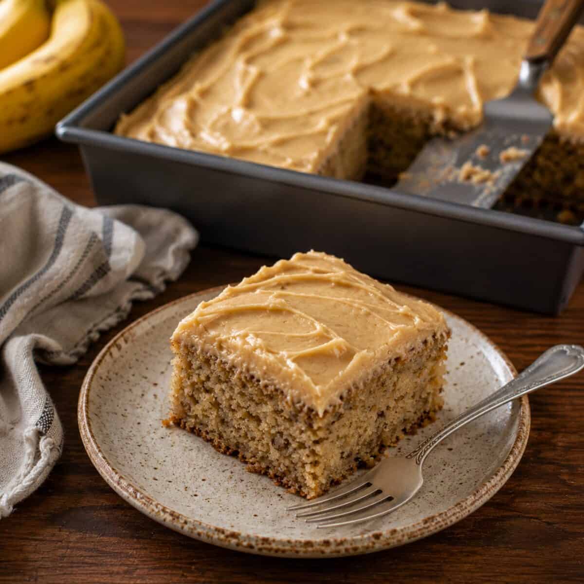 Piece of banana cake with peanut butter frosting on small plate with a fork and the cake pan with spatula in the background.