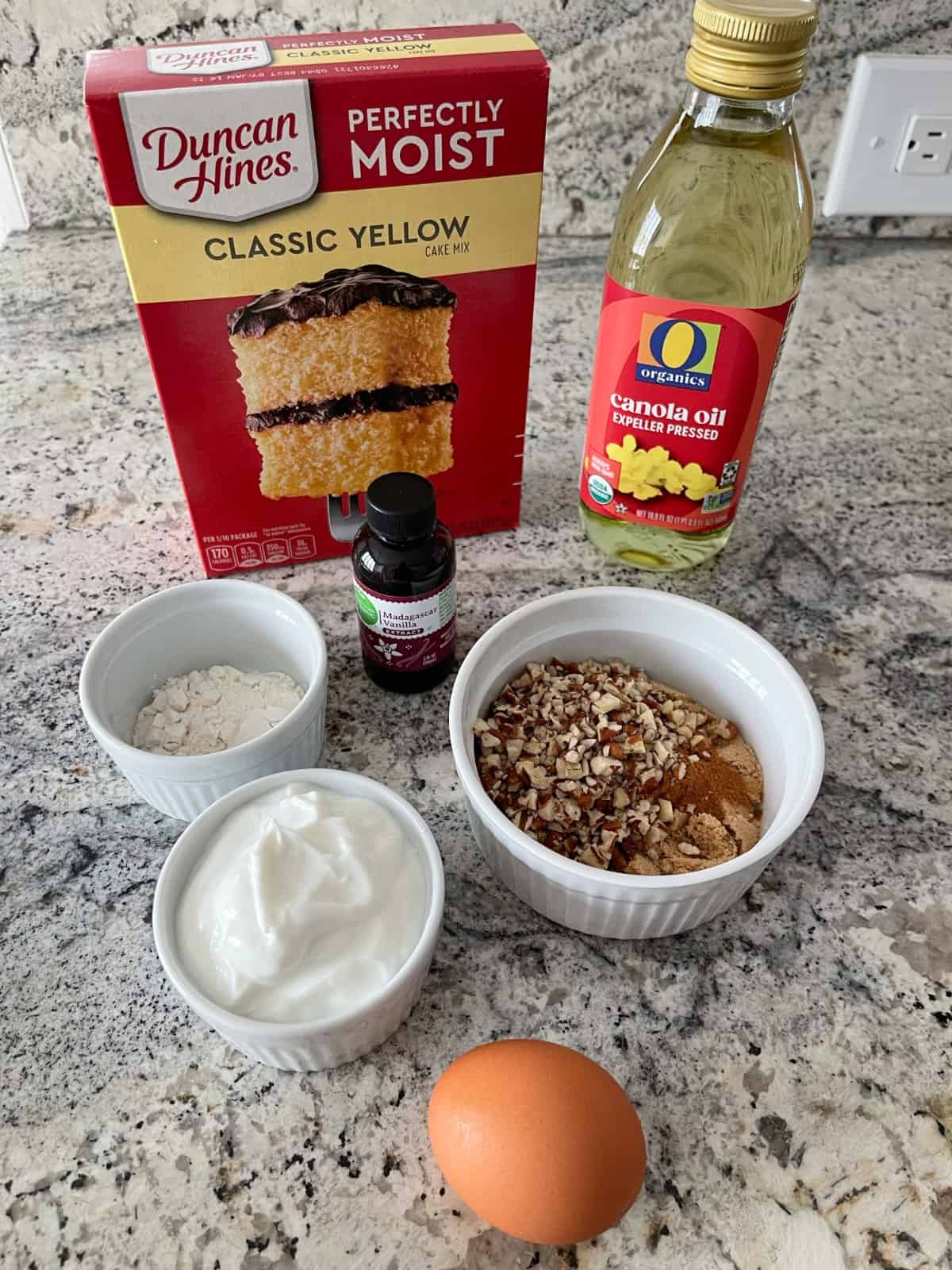 Ingredients including box of Duncan Hines perfectly moist classic yellow cake mix, canola oil, vanilla extract, all-purpose flour, plain Greek yogurt, chopped pecans, cinnamon, brown sugar and an egg.