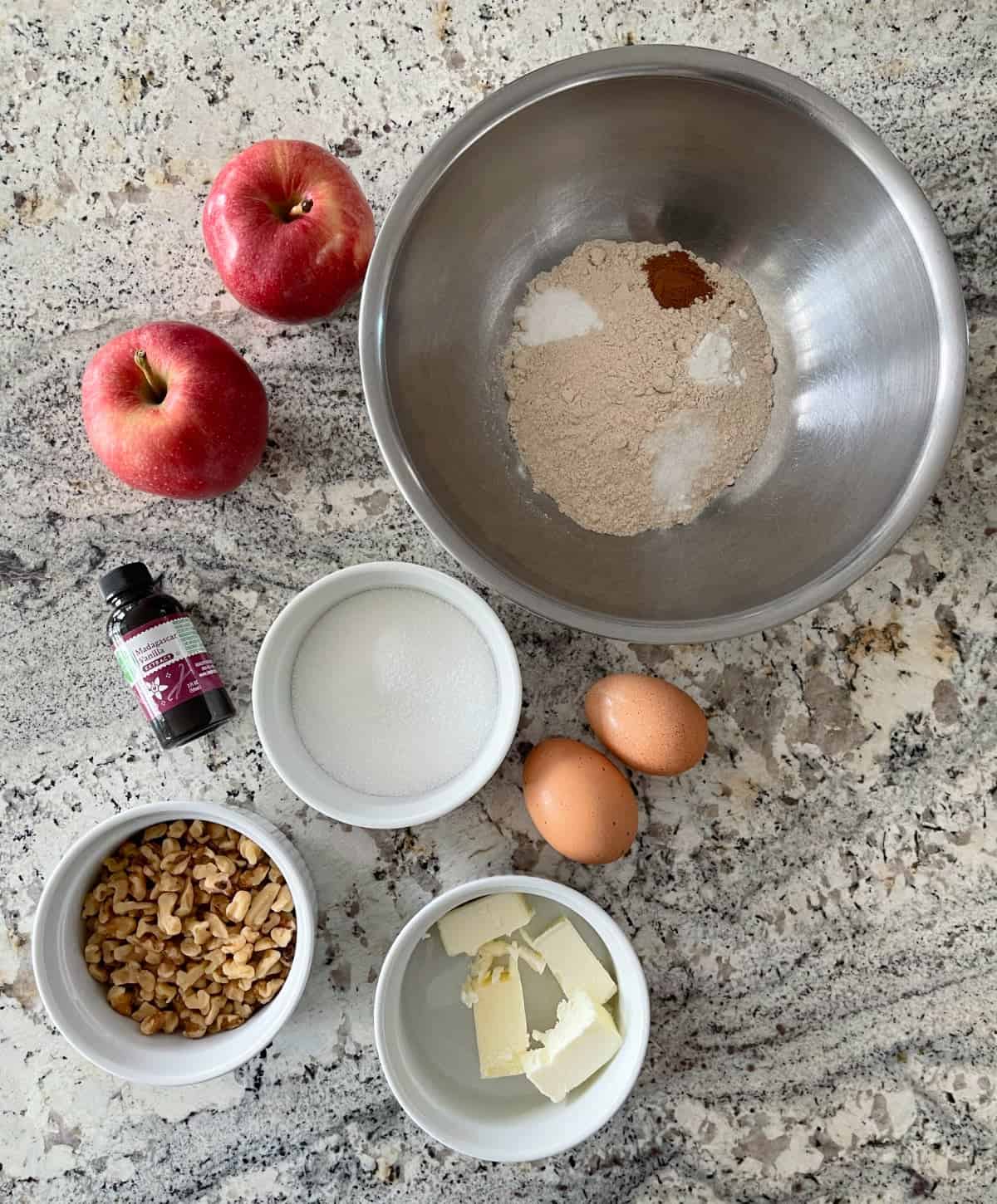 Ingredients including two apples, vanilla extract, chopped walnuts, butter, sugar, two eggs, and a mixing bowl with flour, baking powder baking soda, cinnamon and salt.
