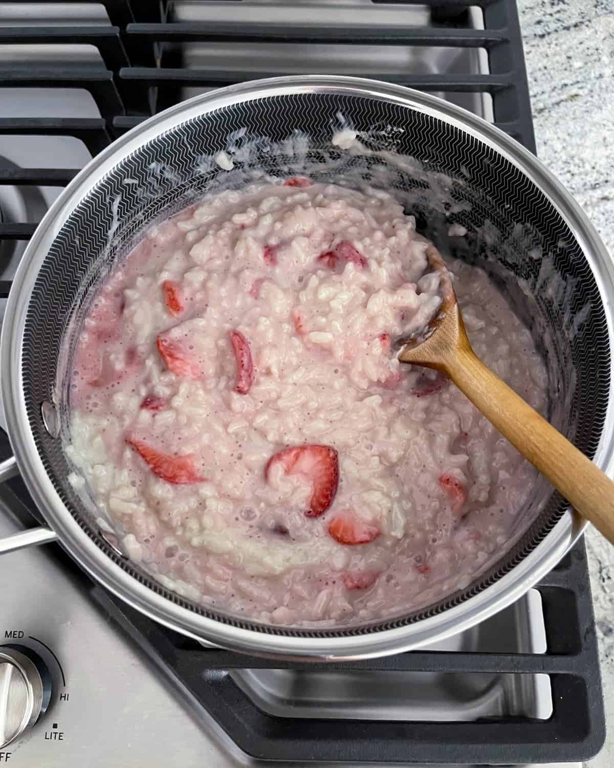 Stirring stovetop strawberry rice pudding in saucepan with wooden spoon.