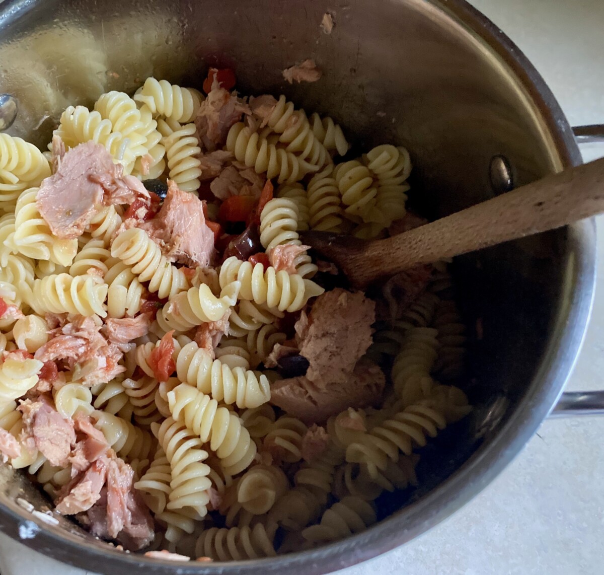 Adding pasta, tuna, olives and capers to pot with large wood spoon.