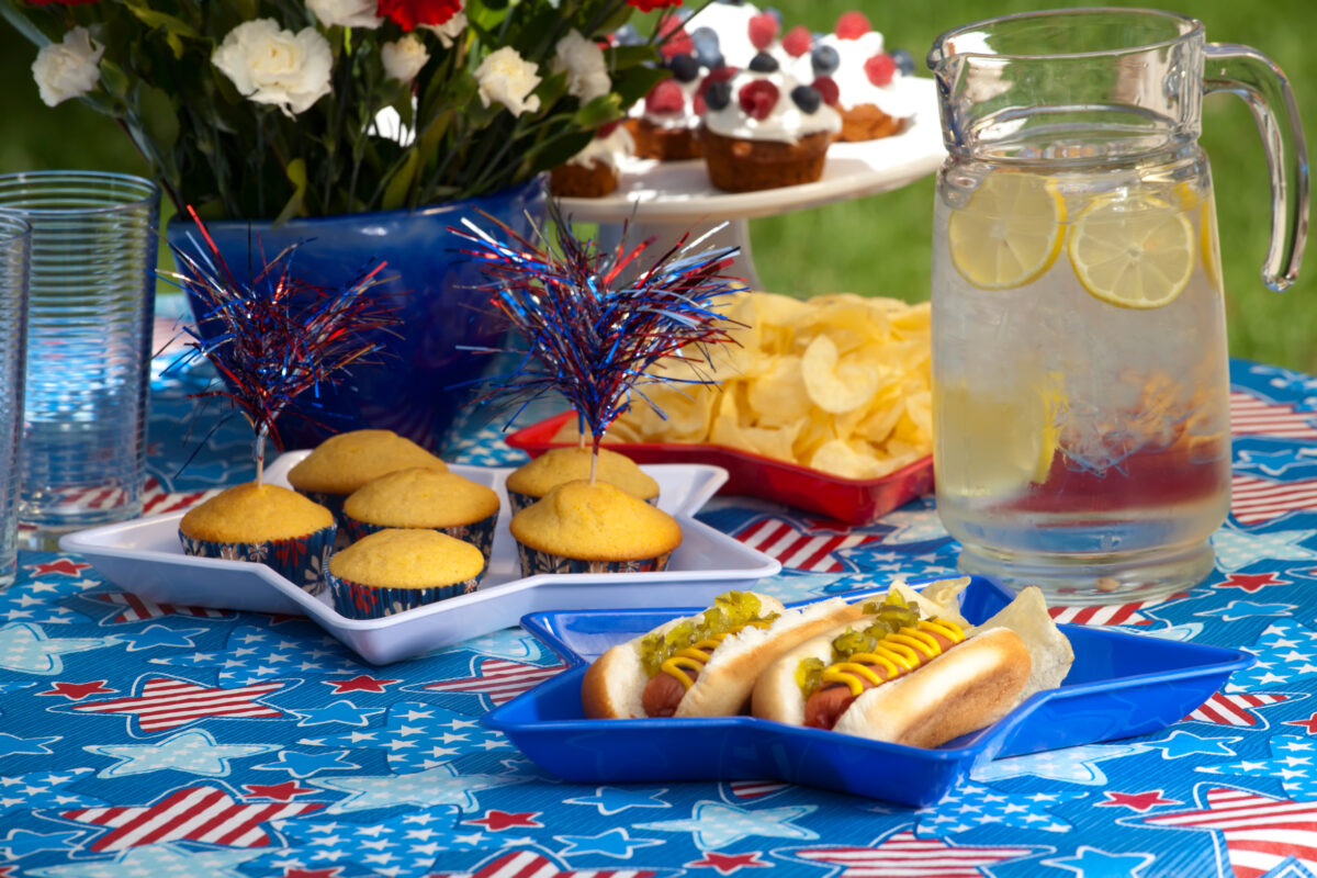 July 4th picnic table decorated in red, white and blue with hot dogs, potato chips and pitcher of lemonade.