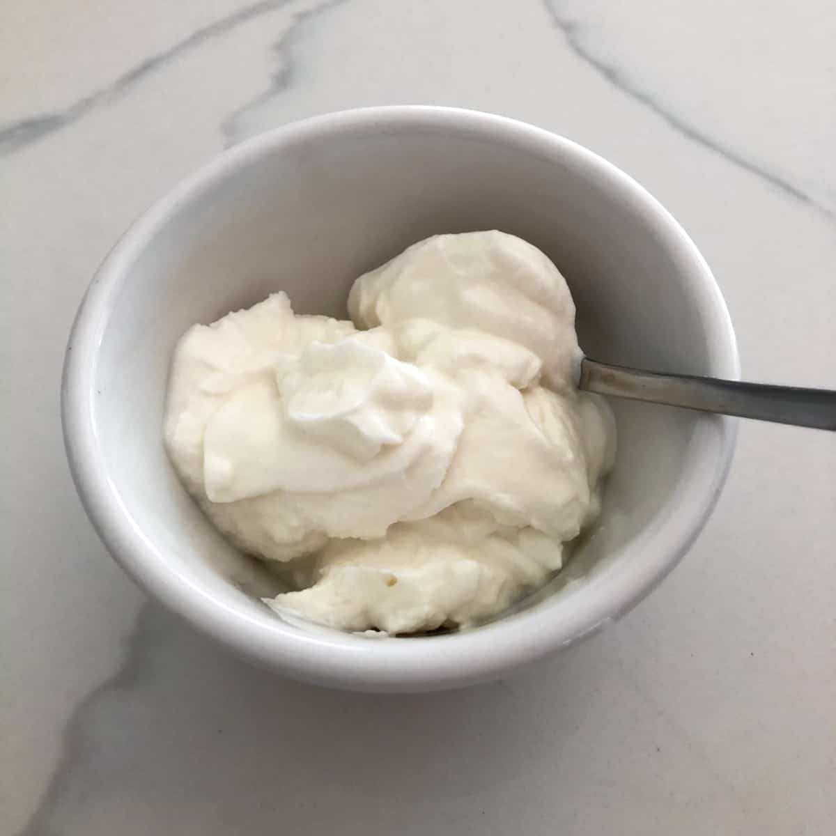 Nonfat Greek yogurt in small white bowl with a spoon on white marble counter.
