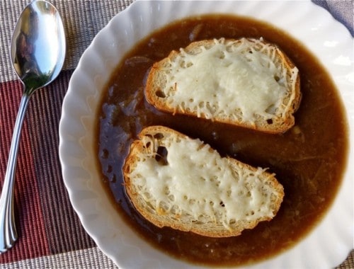 French Onion Soup topped with toasted bread and Swiss cheese in white bowl with spoon.