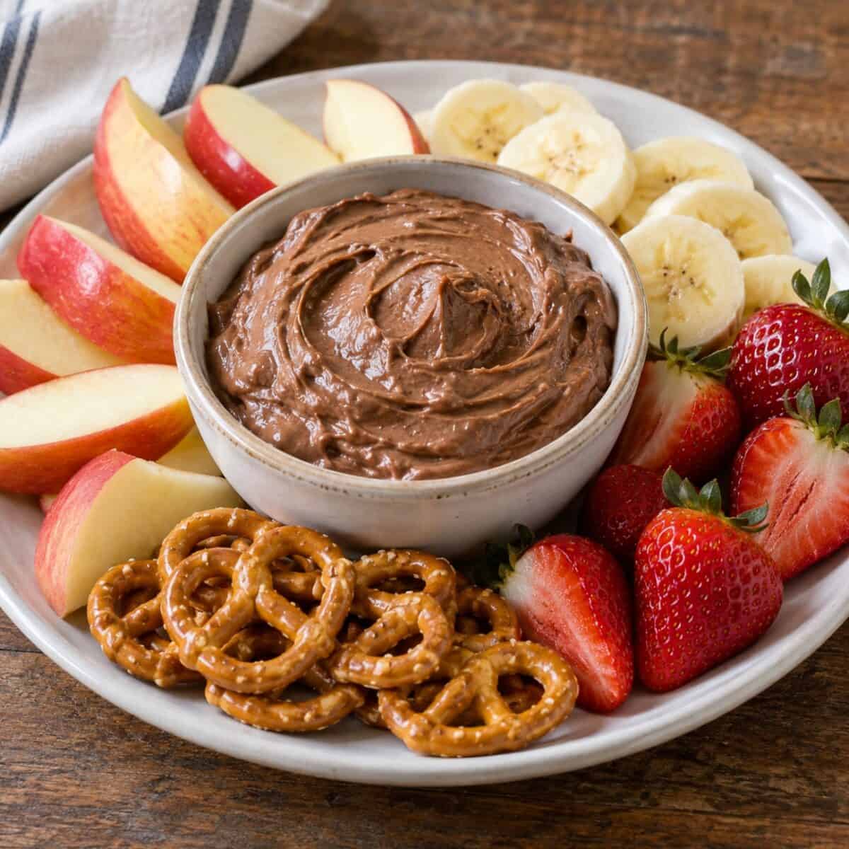 Creamy chocolate peanut butter fruit dip in a bowl with apple slices, banana slices, fresh strawberries and mini pretzels on a serving platter.