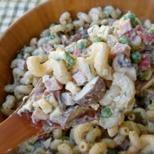close up of ham pasta salad in a wooden salad bowl sitting on a green and white checked tablecloth with serving spoon close up