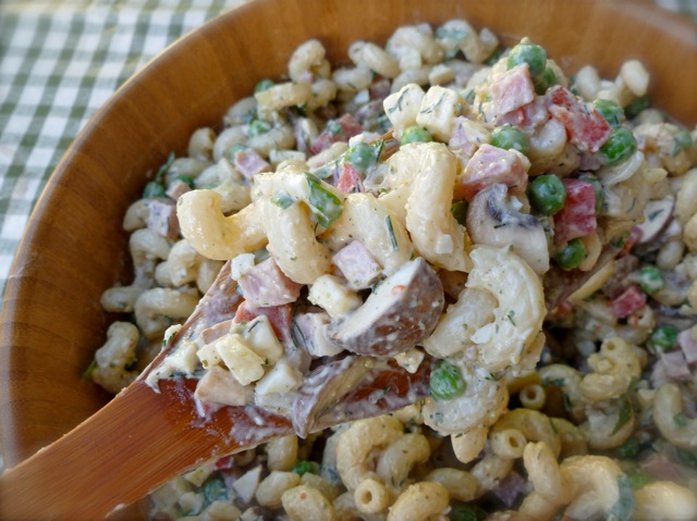 close up of ham pasta salad in a wooden salad bowl sitting on a green and white checked tablecloth with serving spoon close up