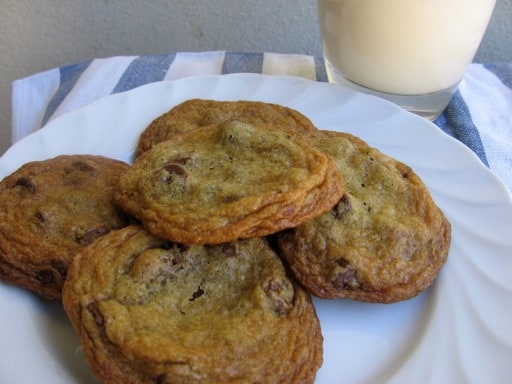 Homemade chewy chocolate chip cookies on white plate with glass of milk in background.