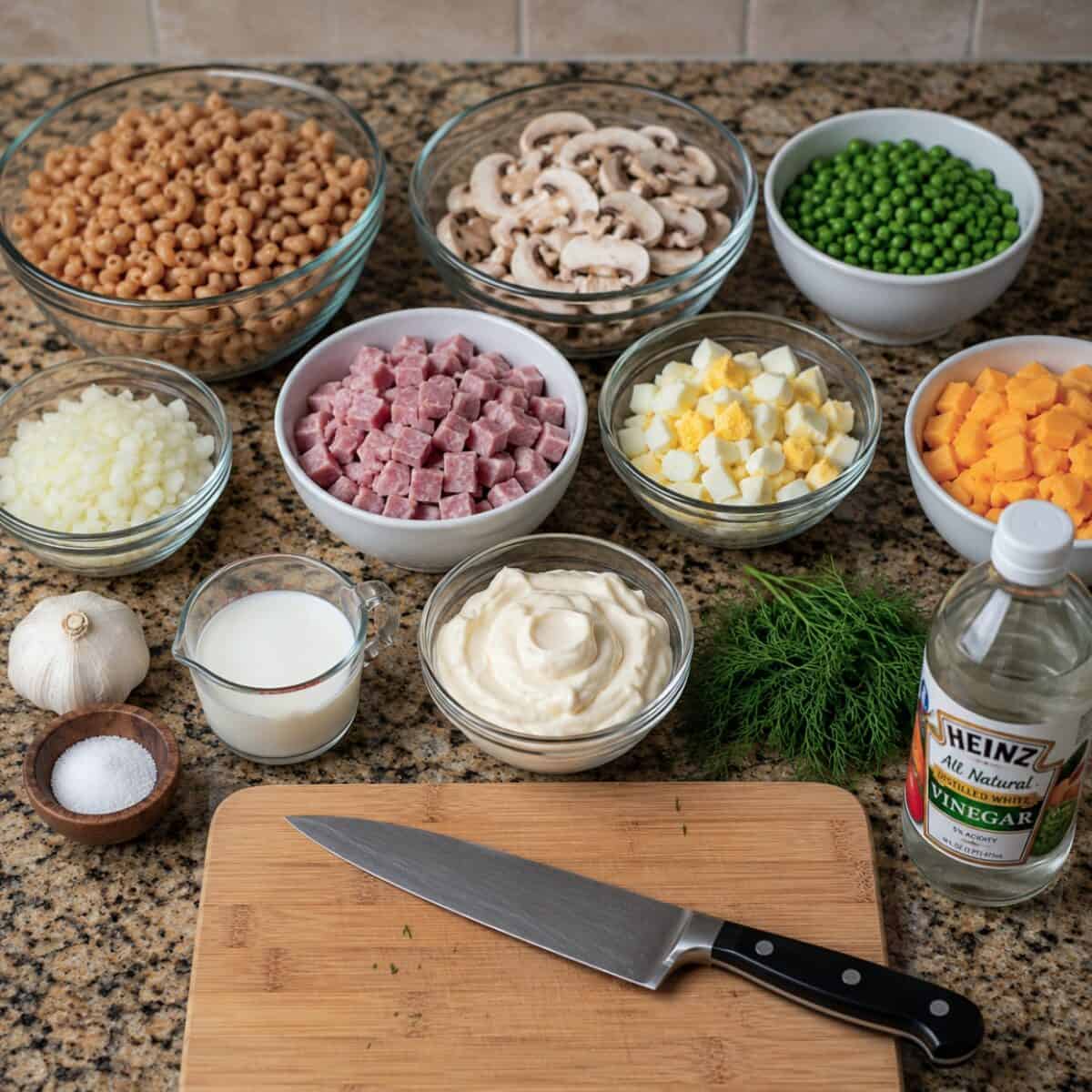 Ingredients including cooked whole wheat elbow macaroni, sliced mushrooms, frozen peas, chopped sweet onion, lean ham cubed, diced hardboiled eggs, diced cheddar cheese, garlic, salt, buttermilk, reduced-fat mayonnaise, fresh dill and distilled white vinegar on kitchen counter.
