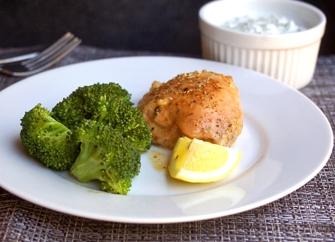 Simple baked lemon chicken thigh on white plate with steamed broccoli florets and fresh lemon wedge.