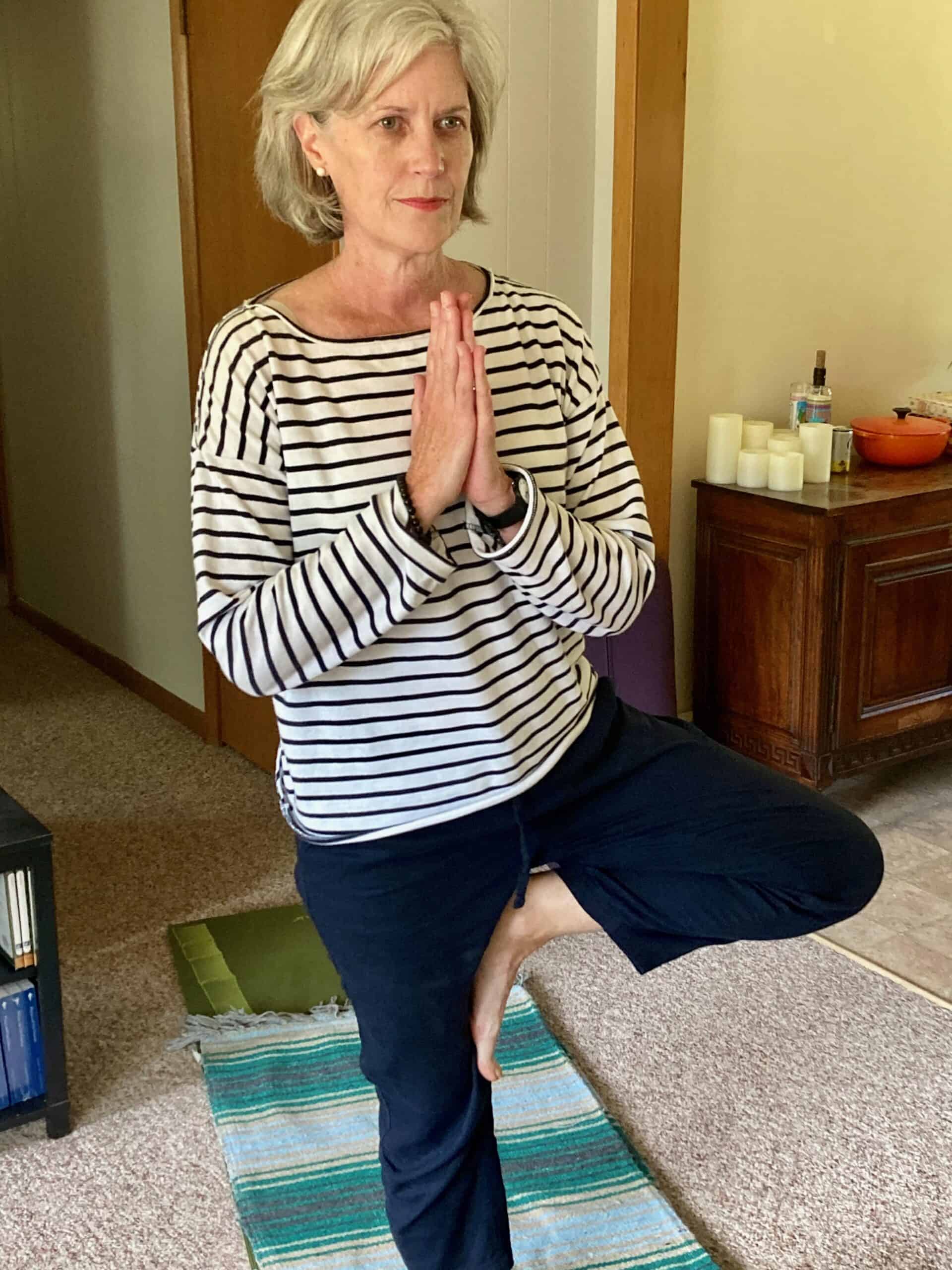 Mature woman gray hair wearing striped shirt and loose pants standing on a yoga mat in Tree Pose