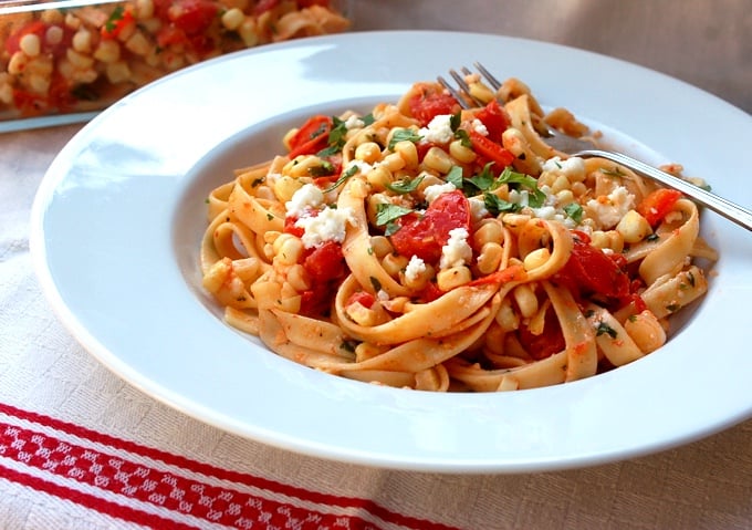 Fettuccine with fresh corn and tomatoes garnished with crumbled cheese and fresh herbs in white bowl with fork.