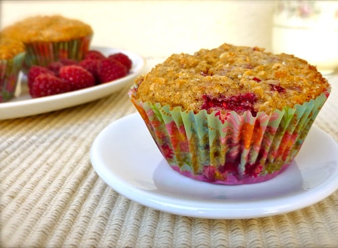Raspberry oatmeal muffin on small white plate with more muffins in the background with fresh raspberries.