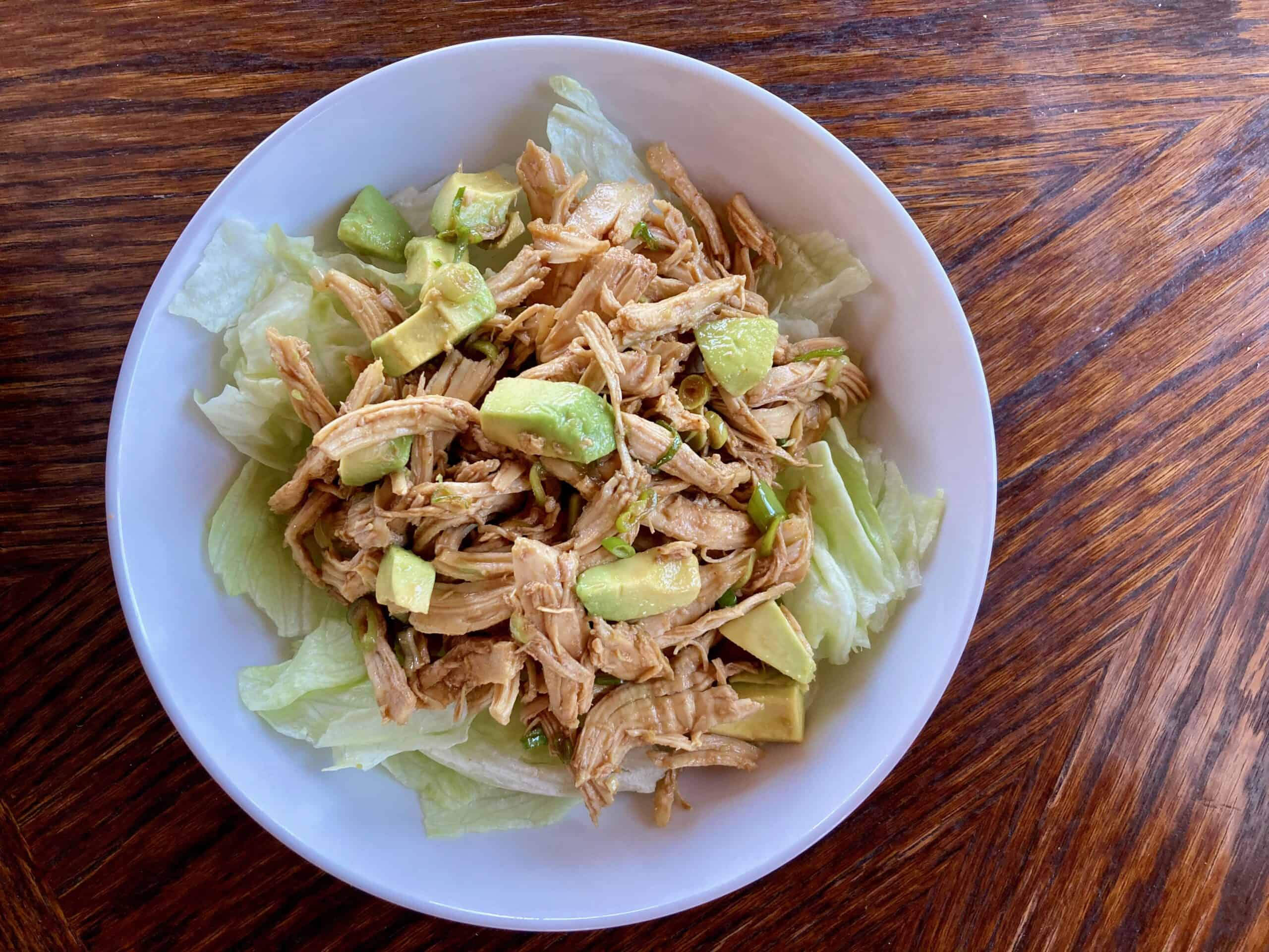 Shredded chicken and avocado on salad greens with Asian flavored dressing on a white plate shot from above.