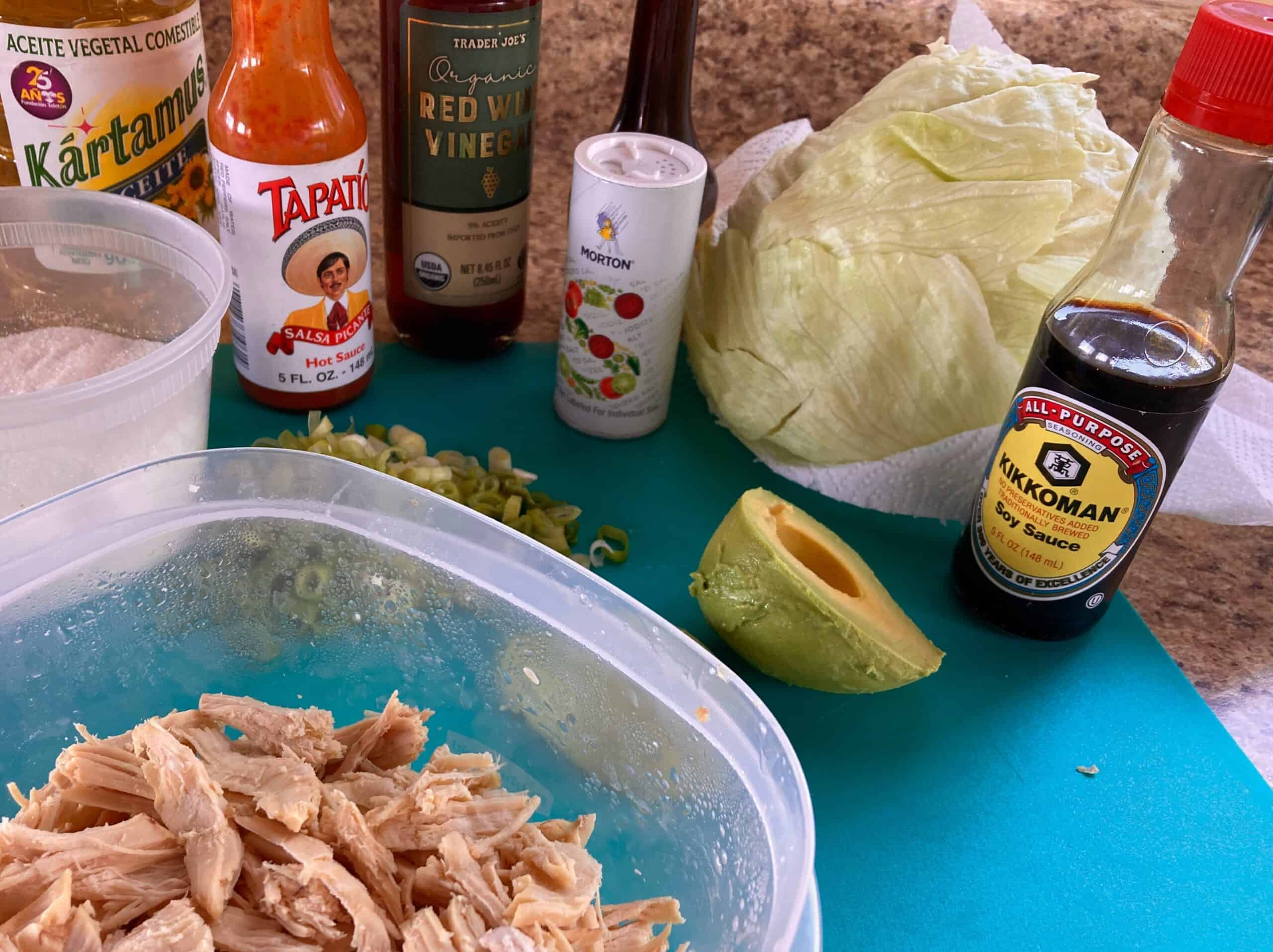 Chicken avocado salad ingredients on counter: shredded chicken, sugar, hot sauce, red wine vinegar, soy sauce, salt, avocado, lettuce.