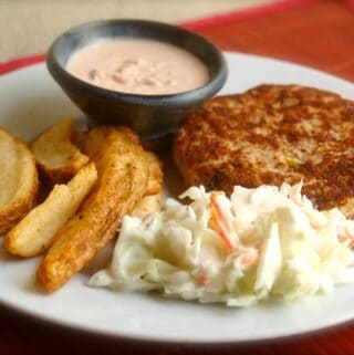 salmon patty on a white plate with coleslaw and potato wedges and a ramekin of sauce on red placemat