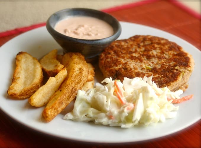 Salmon patty on a white plate with coleslaw and potato wedges and a ramekin of sauce on red placemat.
