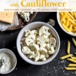 Chopped cauliflower in white bowl, dry penne pasta, cheese, bowl with Greek yogurt and sea salt on black slate for making Cauliflower Macaroni and Cheese.
