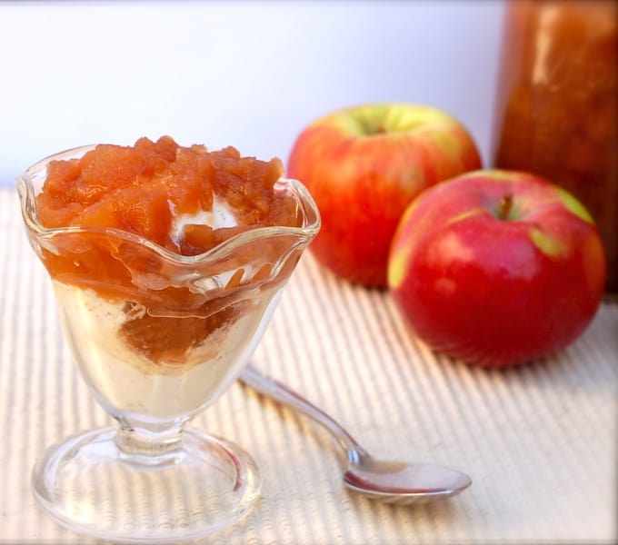 Apple compote over vanilla ice cream in dessert glass with spoon and two fresh apples off to the side.