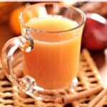 Hot spiced apple cider in glass mug on woven mat with cinnamon sticks, apples and pumpkins in the background.
