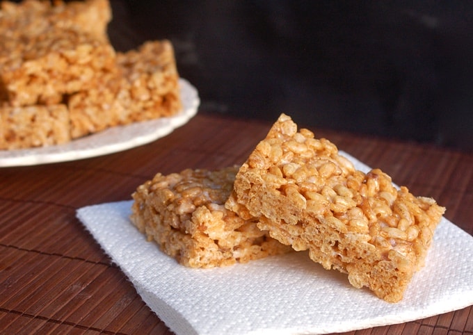 Two Crock Pot Rice Krispie Treats on white napkin with plate of Rice Krispie Treats in the background.