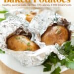Two aluminum foil wrapped baked potatoes with fresh herbs on white plate.