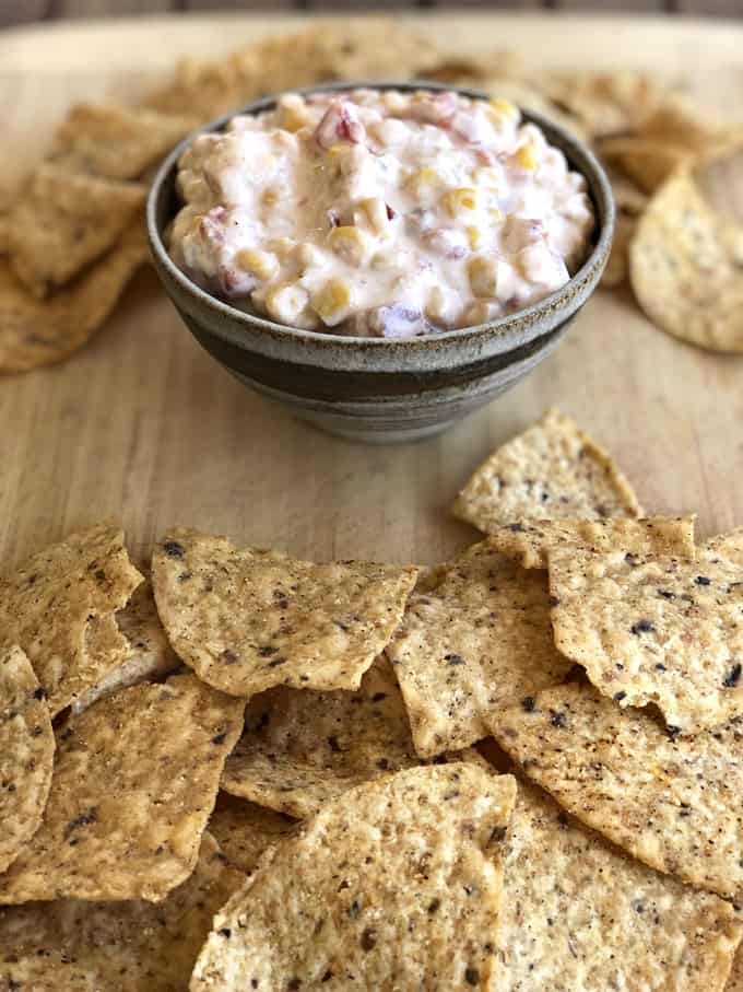 Crock pot corn dip in brown bowl on cutting board with tortilla chips scatter about.