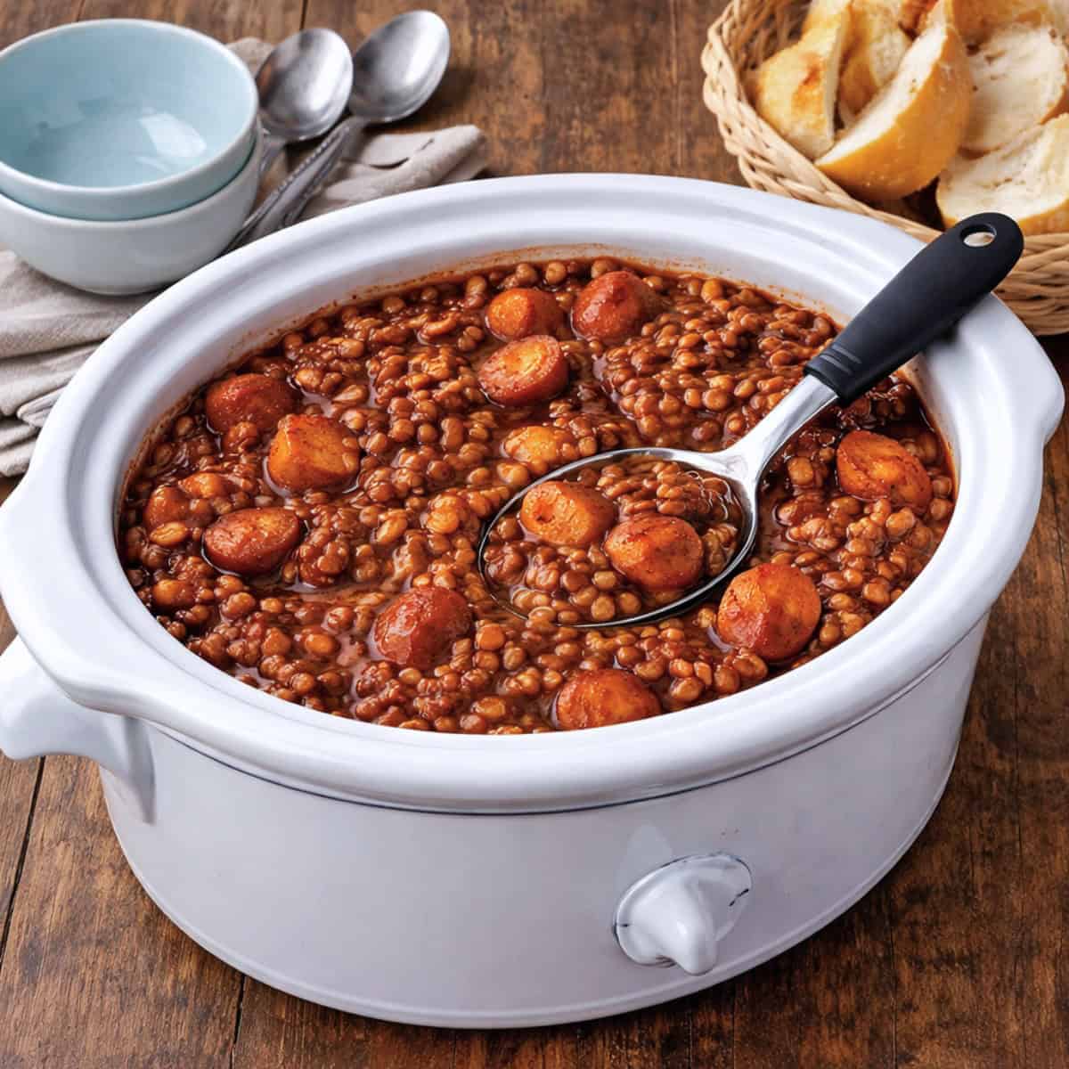 Slow Cooker Barbecue Lentils in white crock pot with serving spoon on wood table with stack of bowls, spoons and basket of crusty bread.