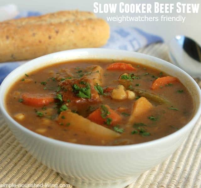 Hearty Slow Cooker Beef Stew garnished with chopped parsley in white bowl with baguette in the background.