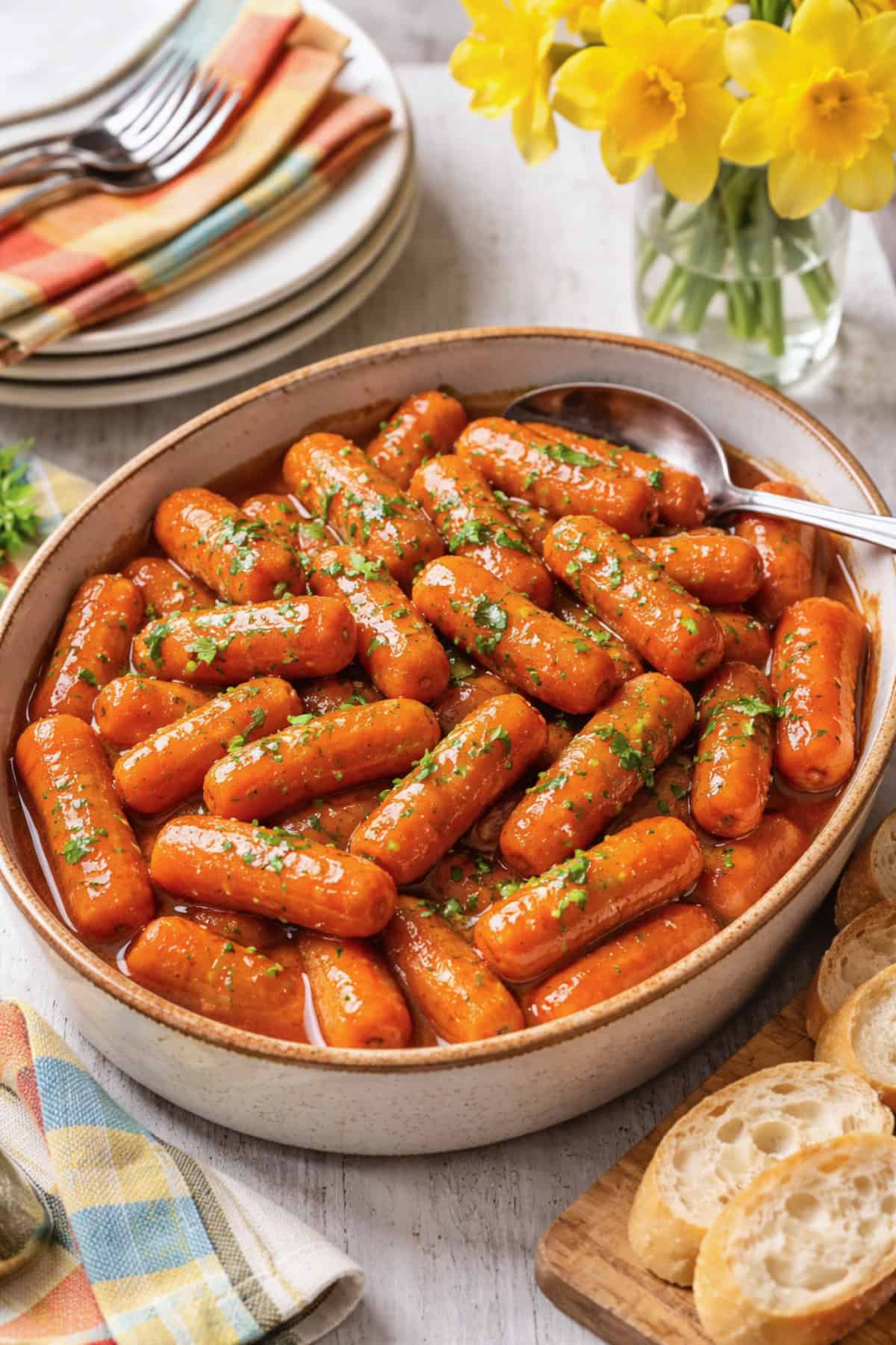 Slow Cooker Apple Glazed Carrots in serving dish on table with sliced baguette and daffodils in a glass vase.