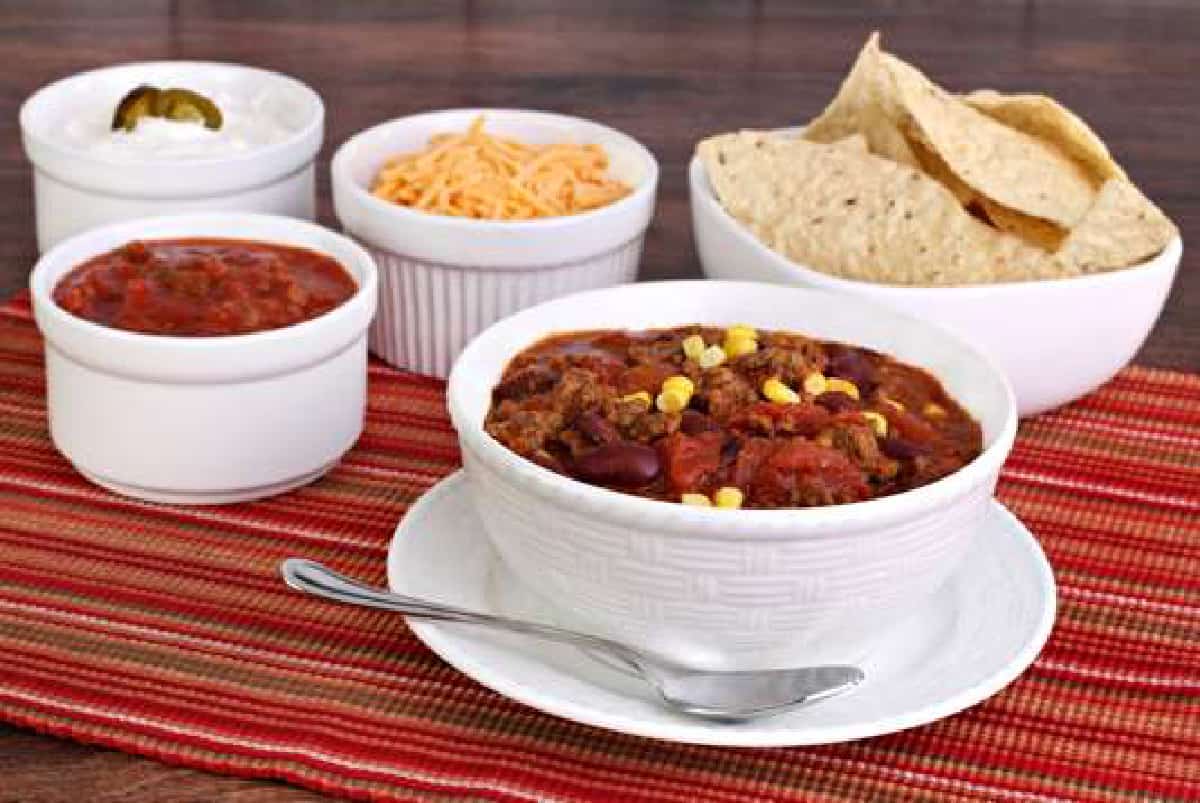 Taco soup in white bowl with toppings for shredded cheese, salsa, tortilla chips and sour cream in small with ramekins.