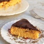 slice of yellow cake topped with thin layer of chocolate frosting on a plate with remaining cake in the background