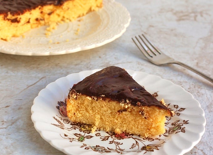 Slice of yellow cake topped with thin layer of chocolate frosting on a plate with remaining cake in the background.