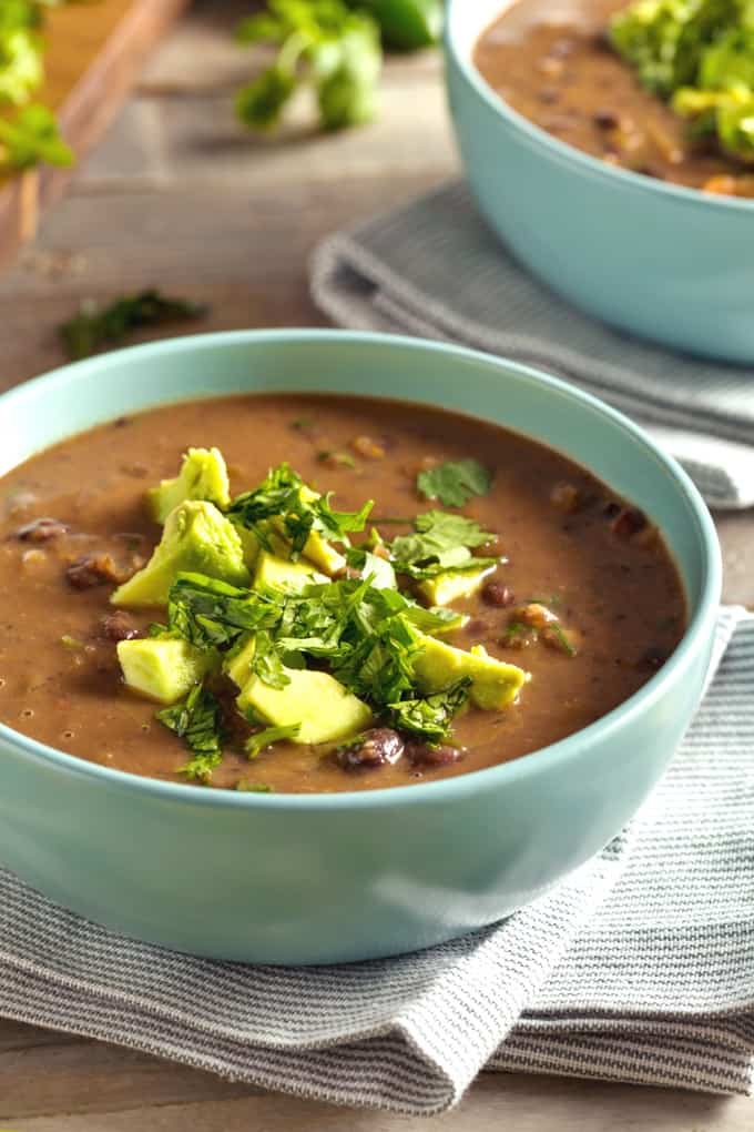 Two green bowls of black bean soup topped with chopped avocado and cilantro.
