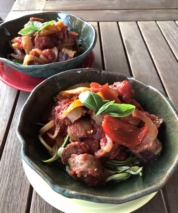 Sausage and peppers over zucchini noodles topped with fresh basil in decorative ceramic bowls.