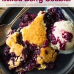 Mixed berry cobbler with a scoop of vanilla ice cream and a spoon on a black plate.