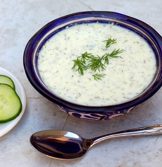 Chilled cucumber soup garnished with fresh dill in blue bowl with spoon and cucumber slices on the side.