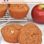 Two apple bran muffins on white napkin with more muffins on cooling rack in the background with a fresh red apple.